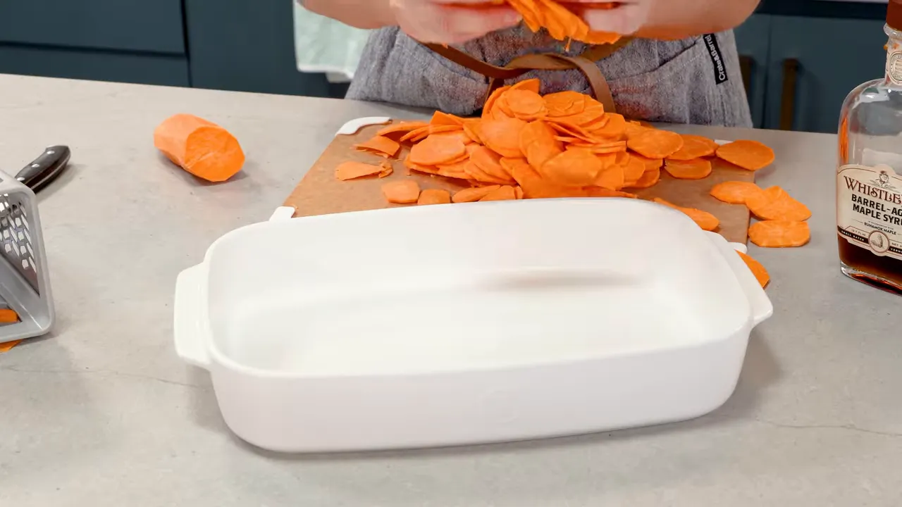 white baking dish on countertop with a pile of thinly sliced sweet potatoes and a bottle of maple syrup nearby
