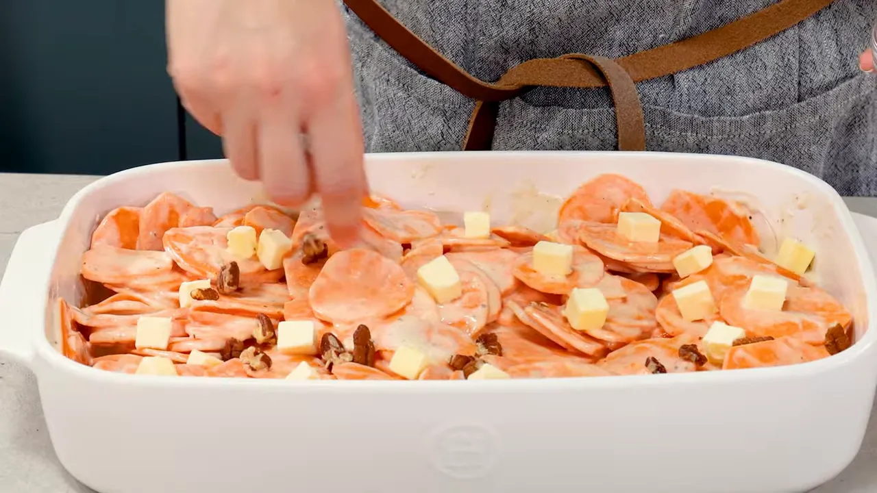 hand sprinkling chopped pecans onto maple-coated sweet potato slices dotted with butter in a white casserole dish