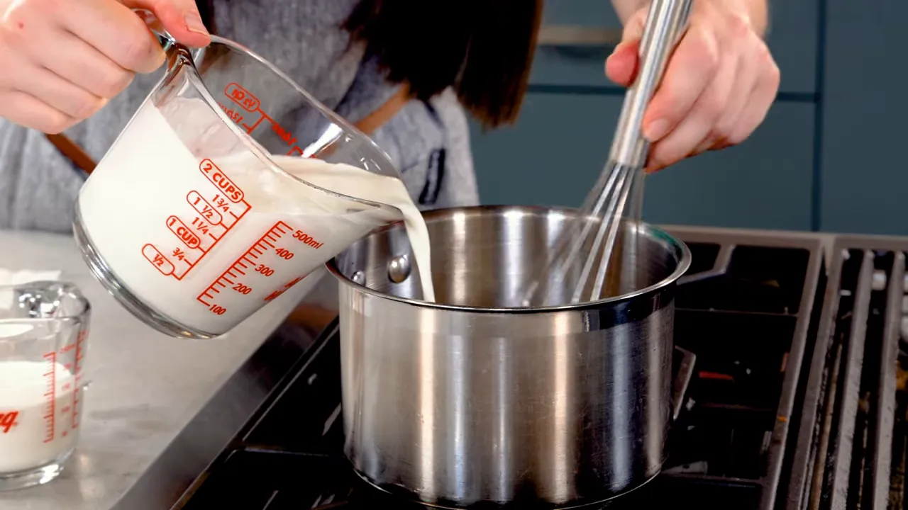 Pouring cream from a measuring cup into a saucepan while whisking to create a smooth sauce
