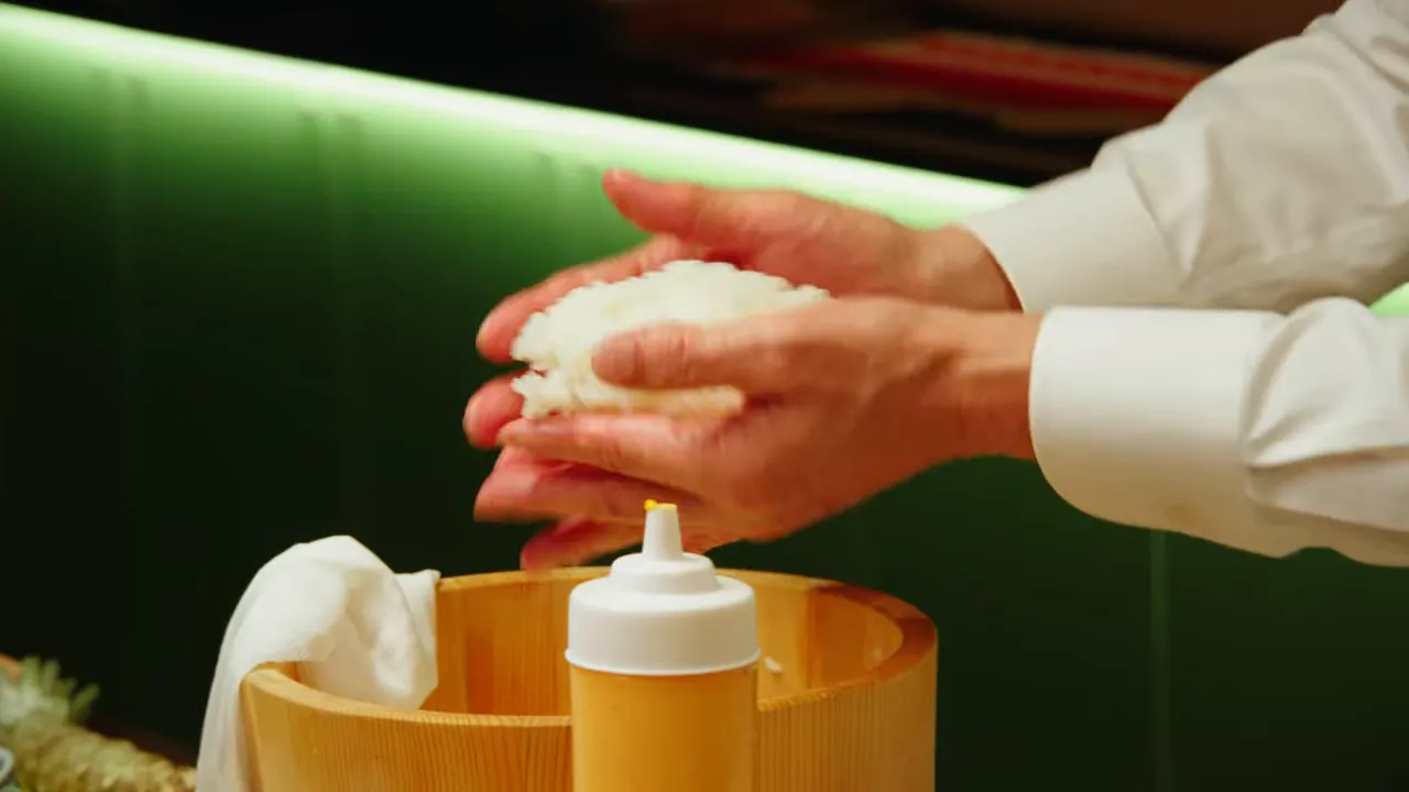 Hands shaping a compact sushi rice ball over a wooden rice tub