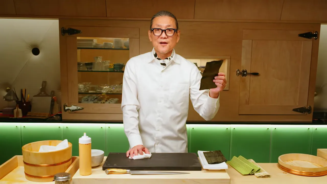 Chef holding a sheet of nori at a sushi counter with tools, rice tub, and knife visible