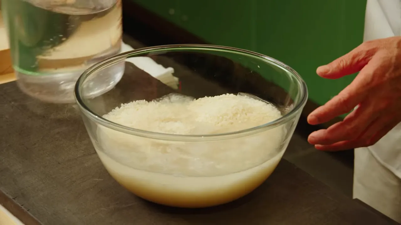 Glass bowl of short-grain rice soaking in water with a measuring jug and a hand nearby