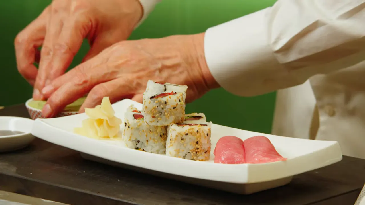 Close-up of a white rectangular plate with sesame-coated inside-out rolls and tuna nigiri while the chef's hands add wasabi or adjust the plate