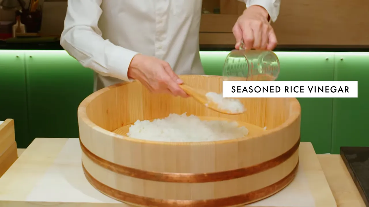 Hands pouring seasoned rice vinegar into a hangiri and folding sushi rice with a wooden paddle