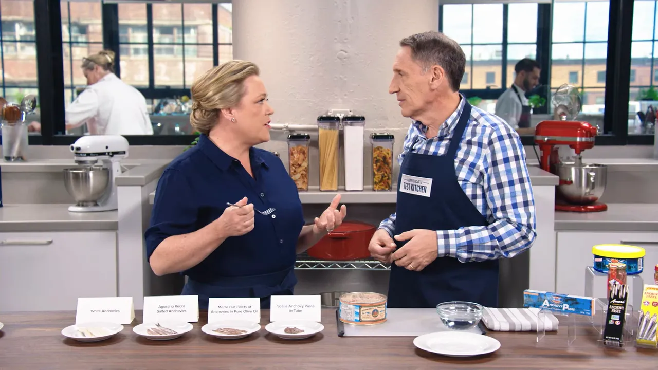 Two hosts at a kitchen counter with plates of different anchovy types and labeled cards laid out in front of them.