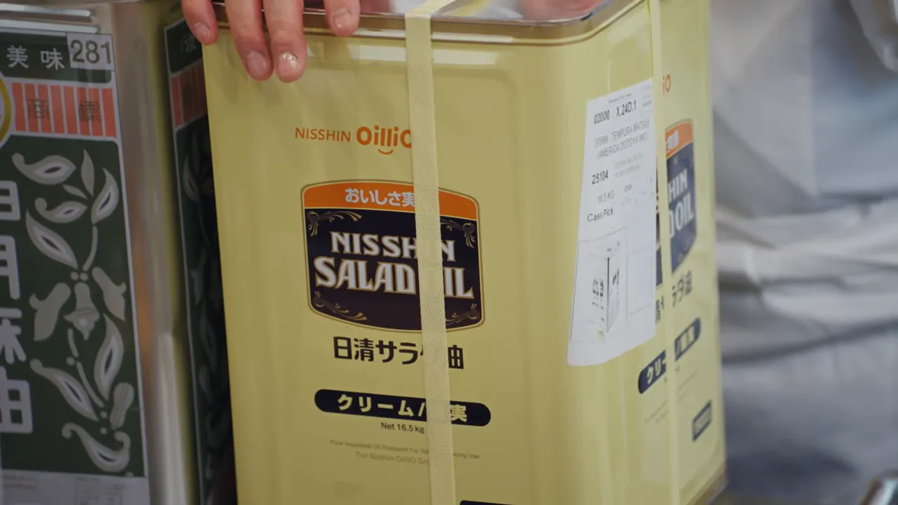 Close-up of a yellow metal 'Nisshin Salad Oil' canister with the label and securing strap clearly visible.