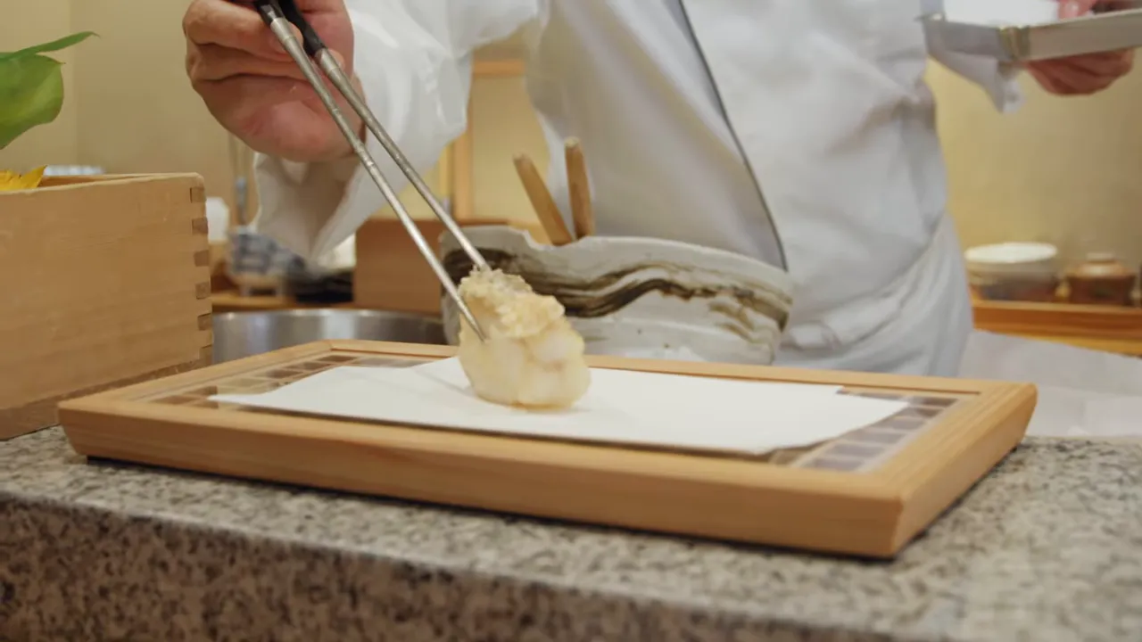 Hands using chopsticks to place a piece of tempura on a paper-lined wooden tray