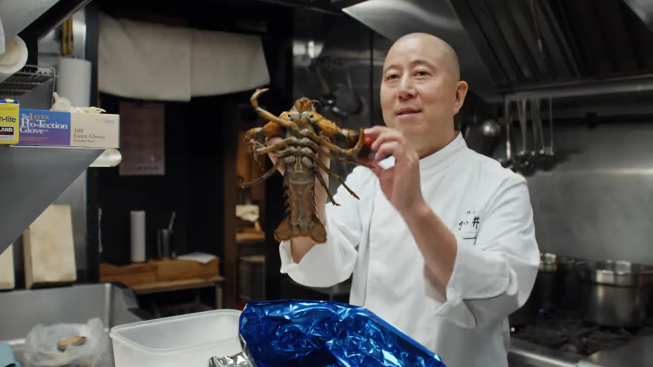 Chef in a kitchen holding a whole lobster over the prep counter.