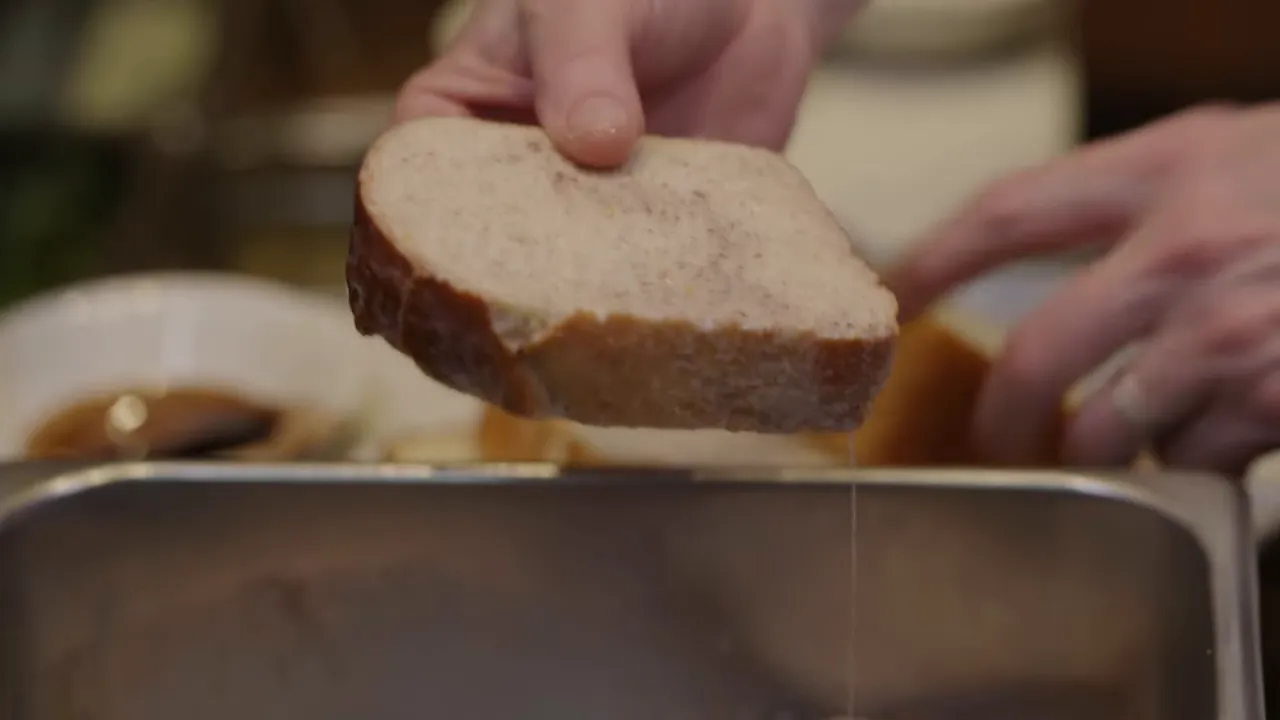 Close-up of a brioche slice lifted above a batter pan with batter dripping from the bread