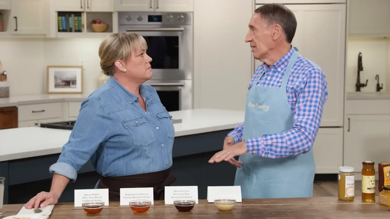 Wide shot of a kitchen counter with four sample bowls of honey, label cards, and jars at the edge of the frame.