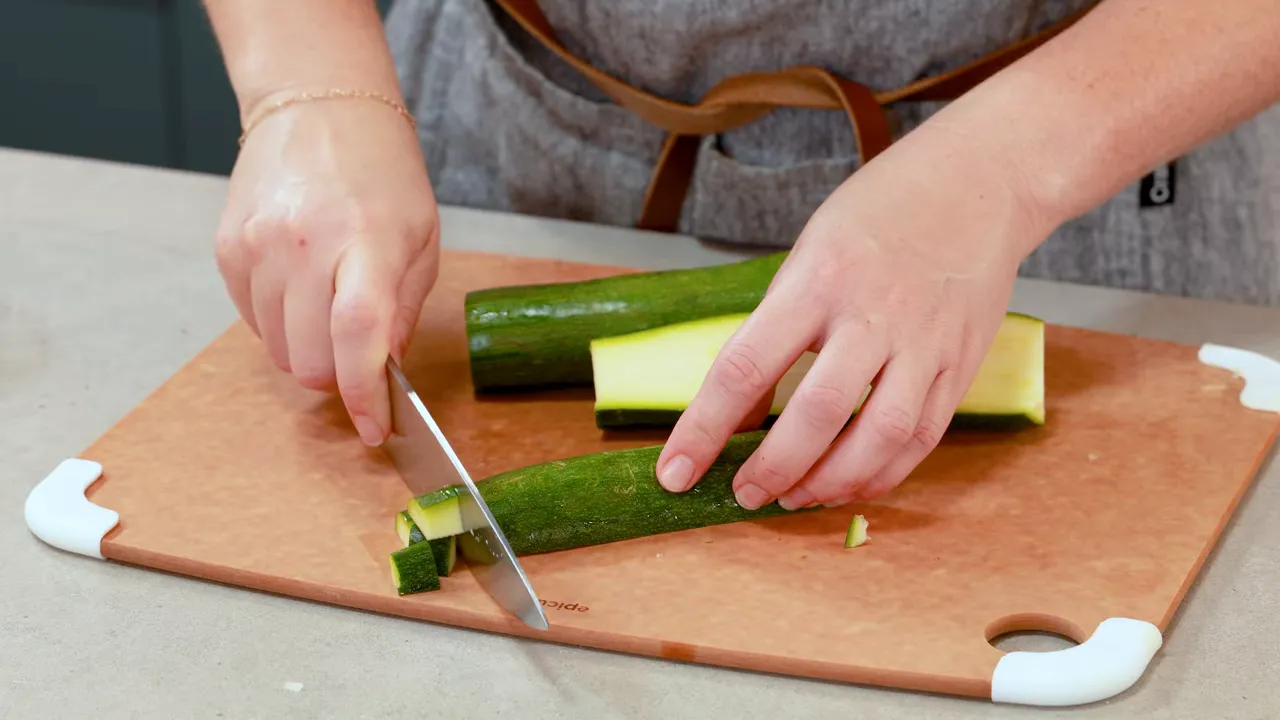 Hands chopping zucchini into small pieces on a cutting board