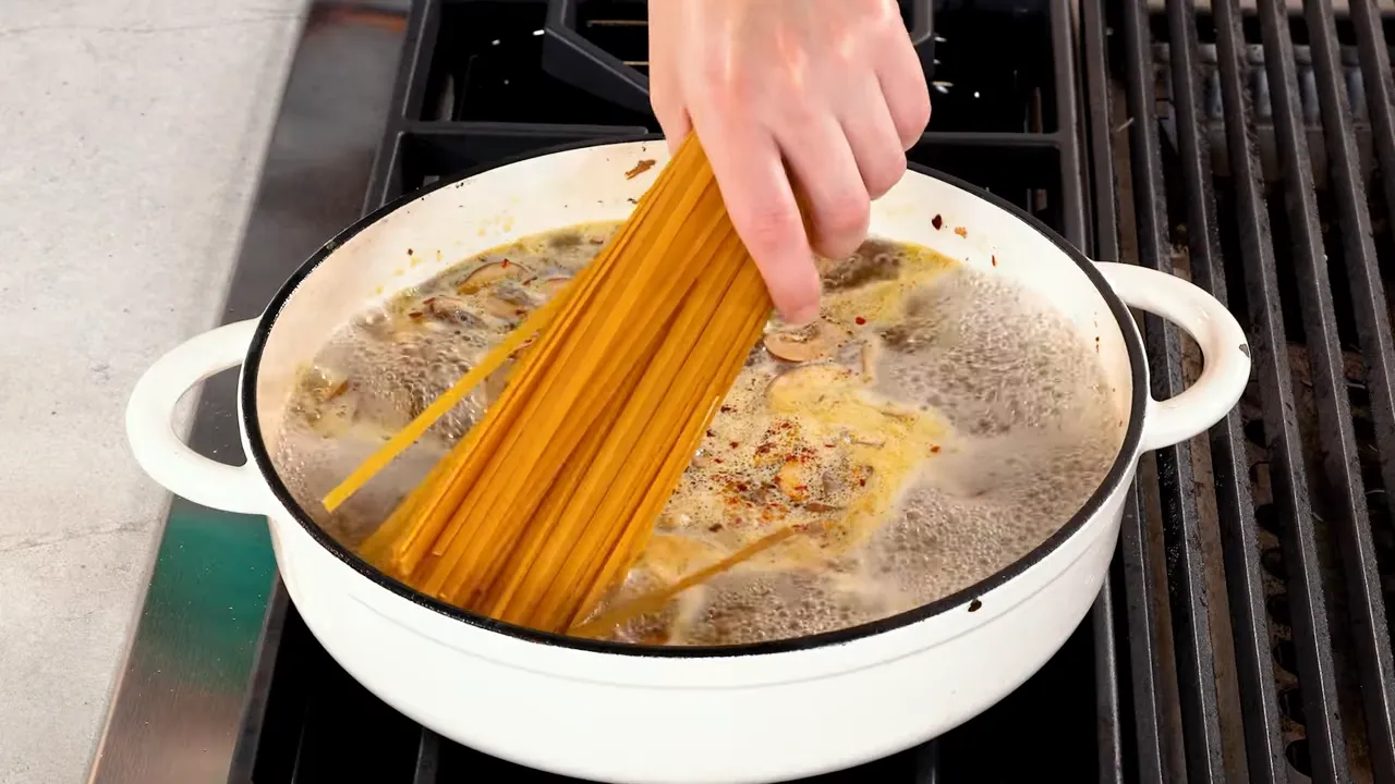 Hand adding long pasta into a boiling pot with mushrooms and broth