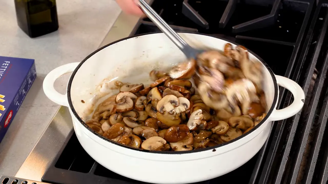 Sliced mushrooms being stirred and sautéed in a white enameled pan on the stovetop.