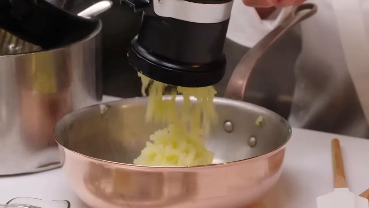 Potatoes being pressed through a potato ricer into a copper pan with steam rising, showing the ricer in action.