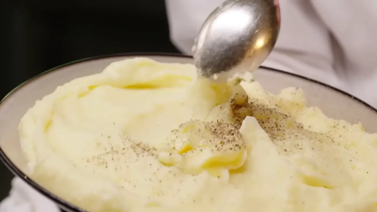 A serving spoon lifting a scoop of silky mashed potatoes from a bowl, showing melted butter and cracked pepper.