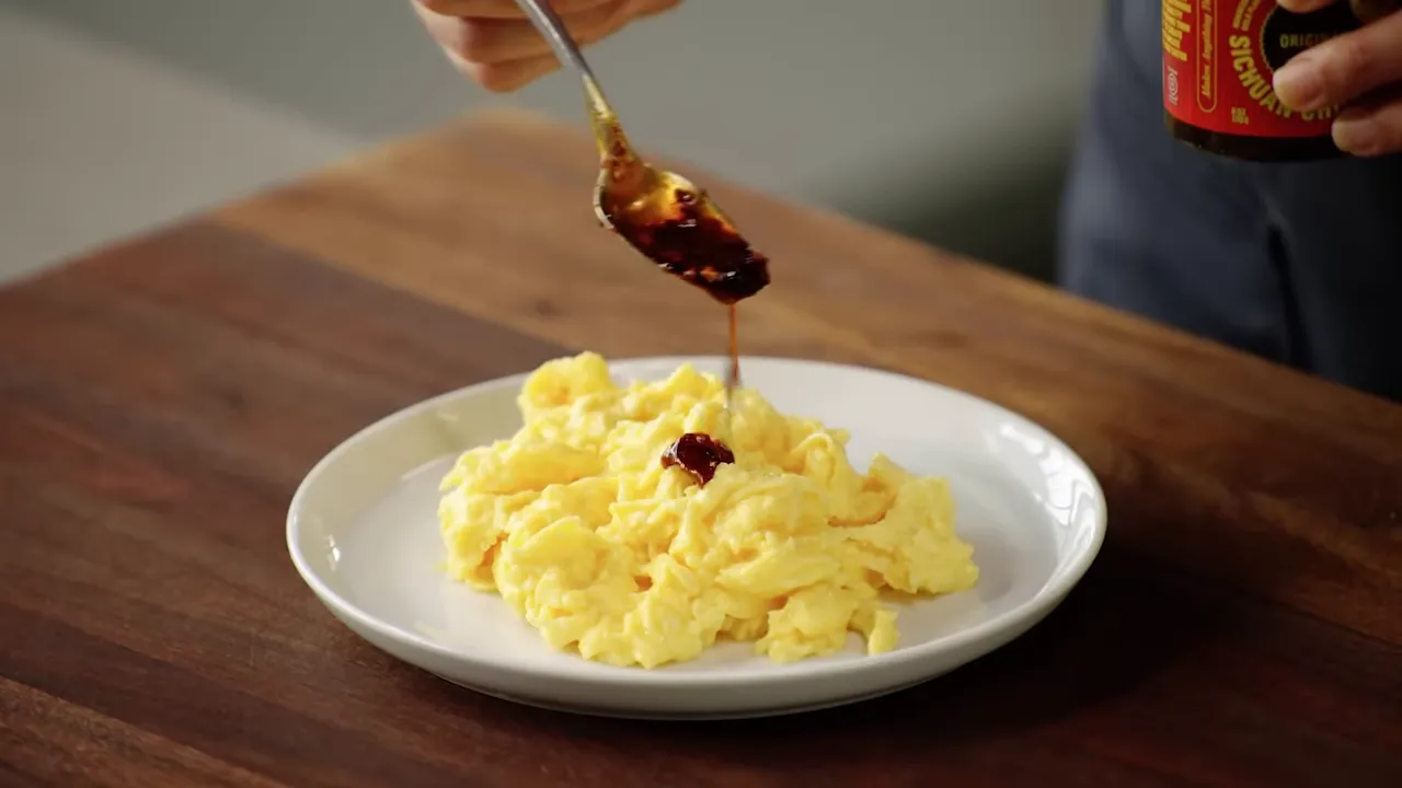 Spoon drizzling chili crisp over scrambled eggs on a white plate.