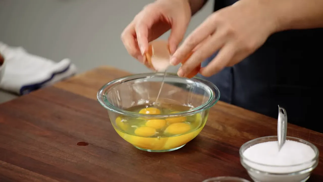 Hands cracking an egg with egg white falling into a glass bowl containing multiple yolks