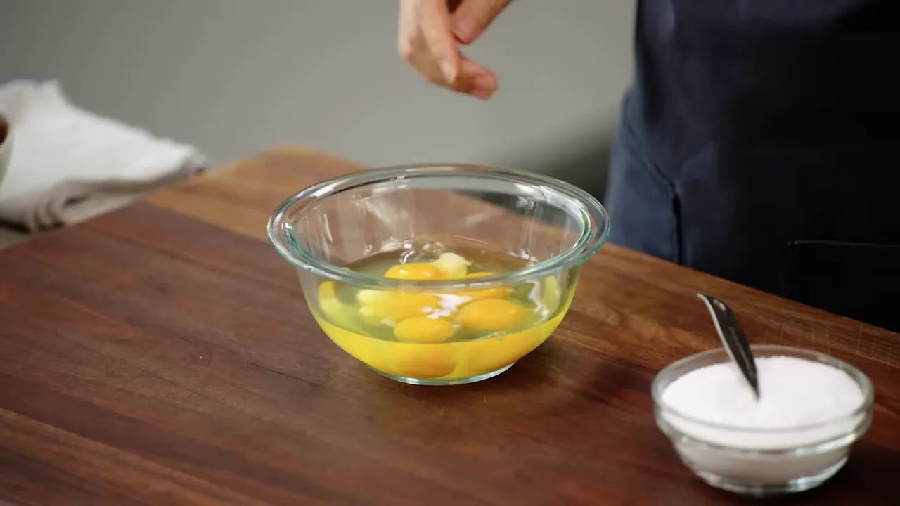 Hand sprinkling salt into cracked eggs in a glass bowl on a cutting board