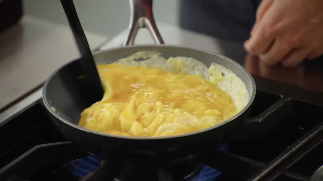 Close-up of scrambled eggs forming curds in a nonstick skillet being folded with a rubber spatula over a blue gas flame