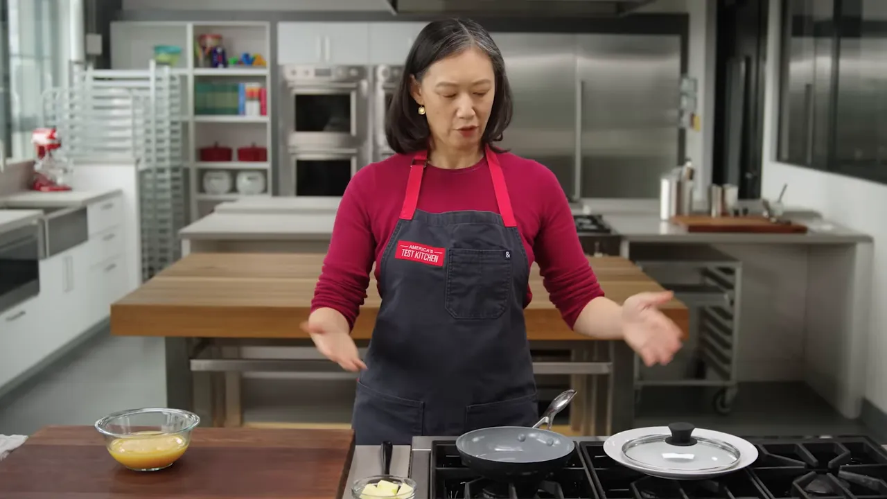 Cook standing behind a prep station with a glass bowl of whisked eggs, a small dish of butter, and a nonstick skillet on a gas stove