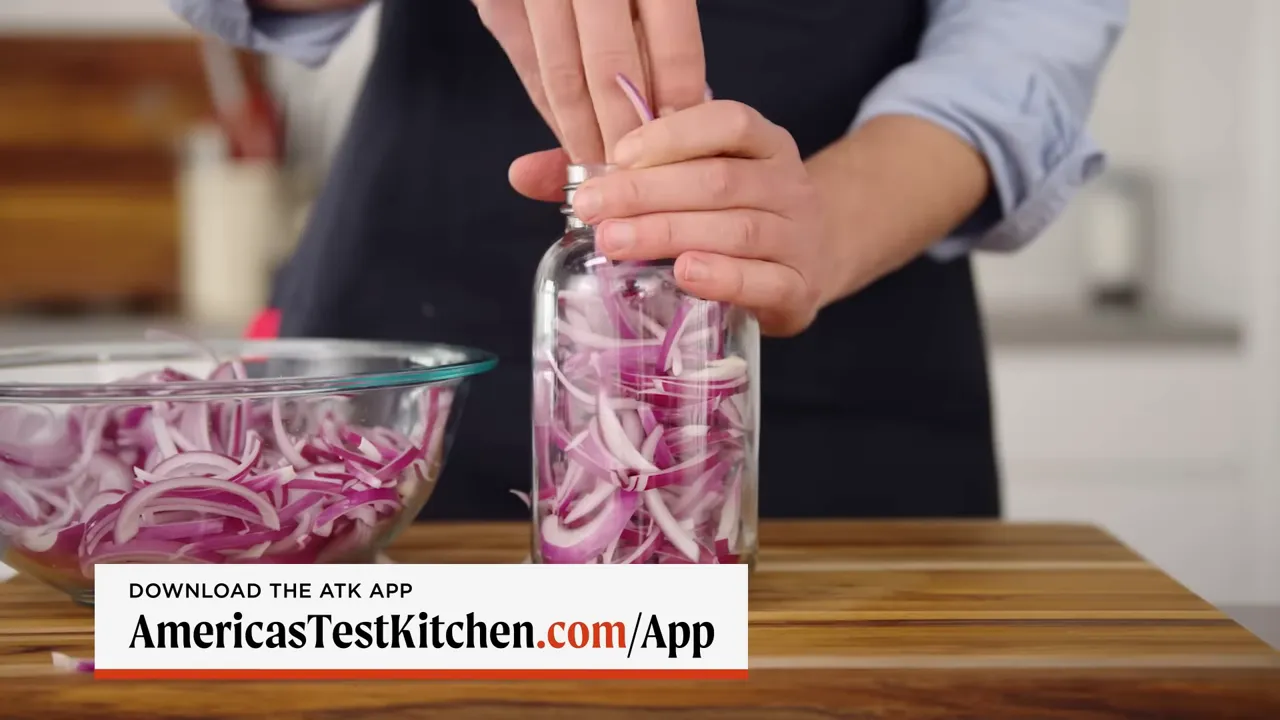 Hands packing thinly sliced red onions into a glass jar with a bowl of slices nearby
