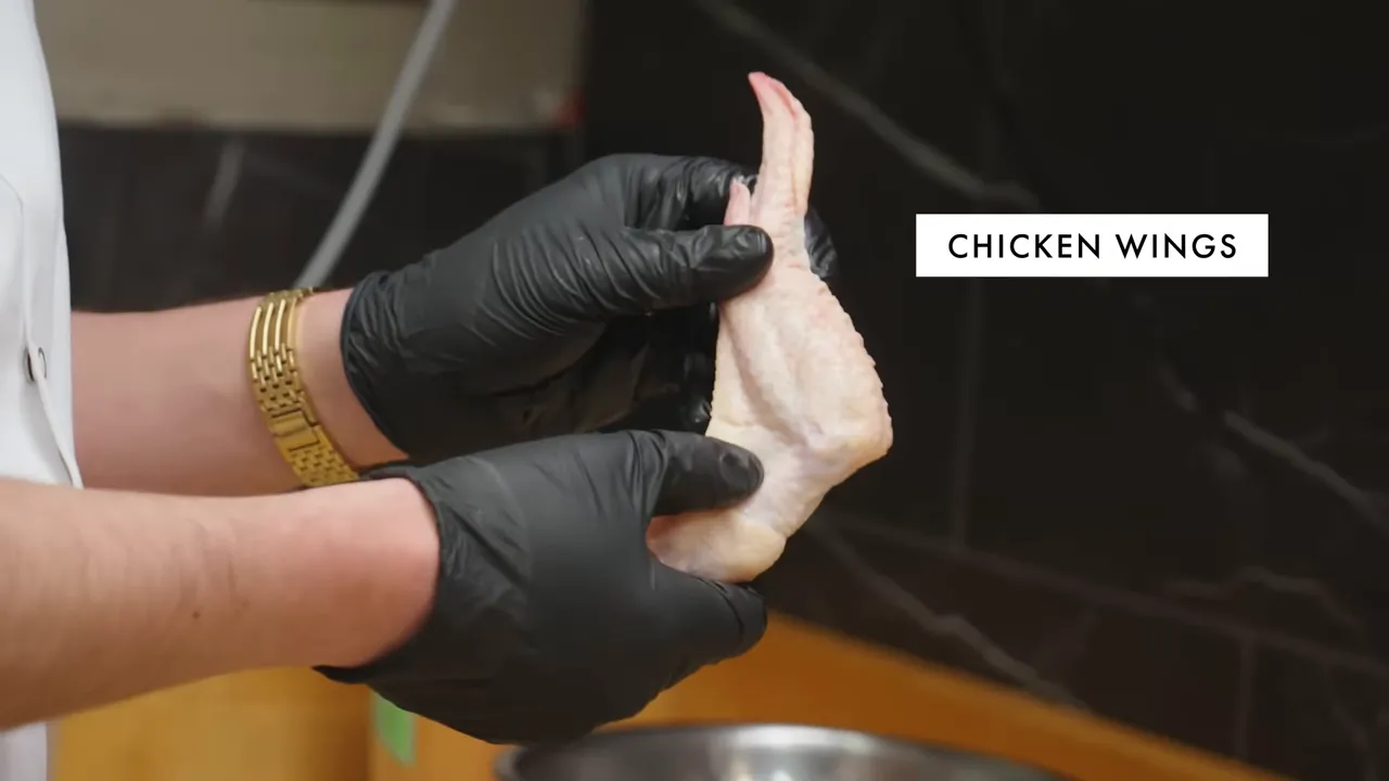 Close-up of gloved hands holding a raw chicken wing, with a 'CHICKEN WINGS' label to the right.