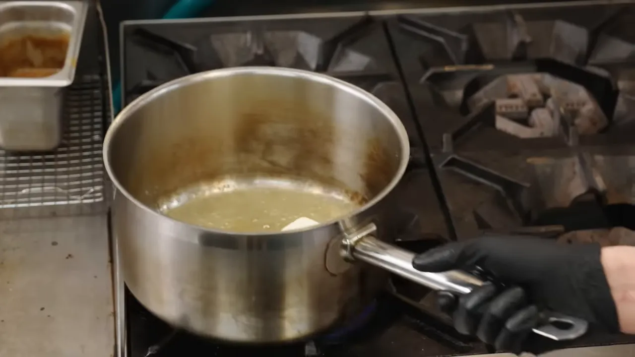 Close-up of a saucepan with a melting chunk of butter and a gloved hand on the handle over a gas flame