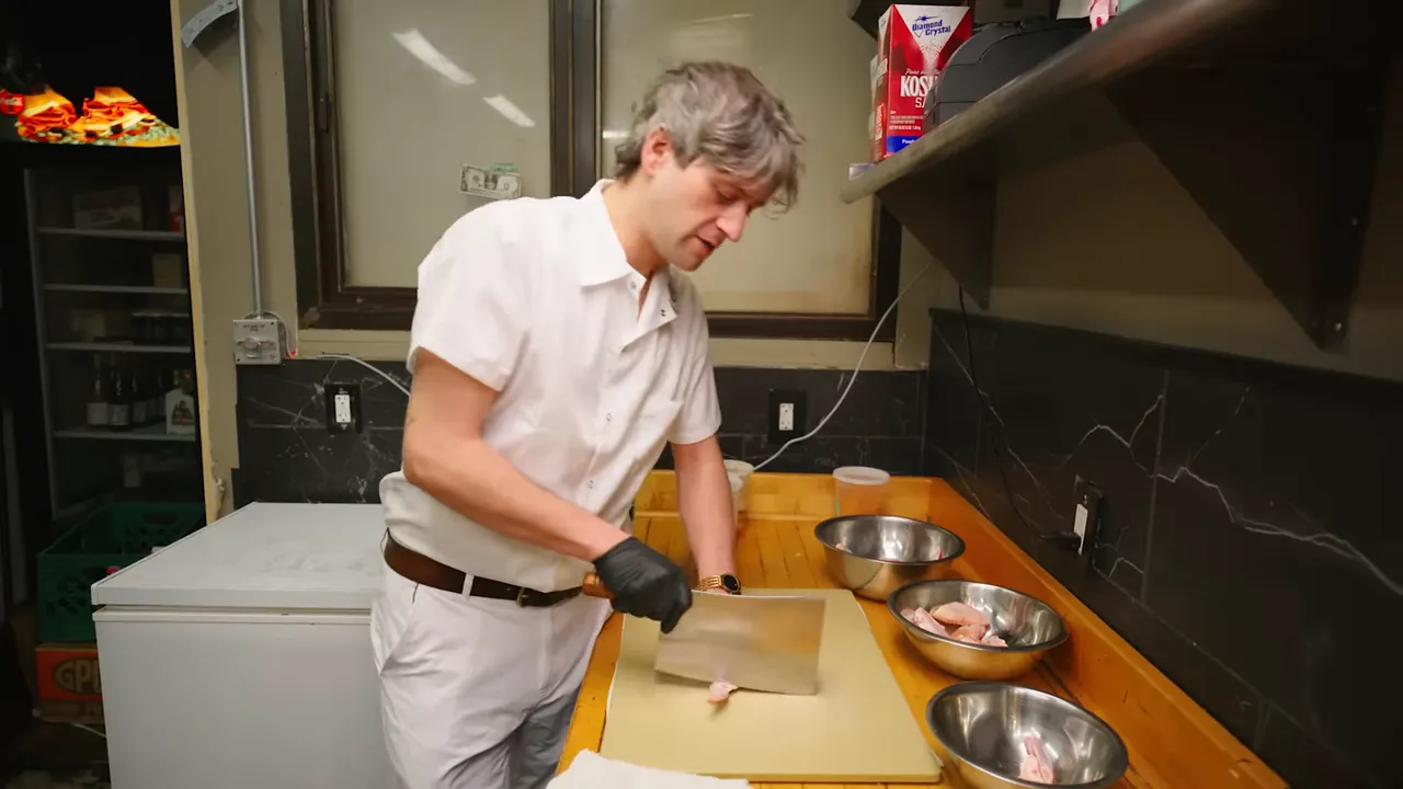 Cook trimming and separating chicken wings on a cutting board with bowls and a cleaver at a prep station