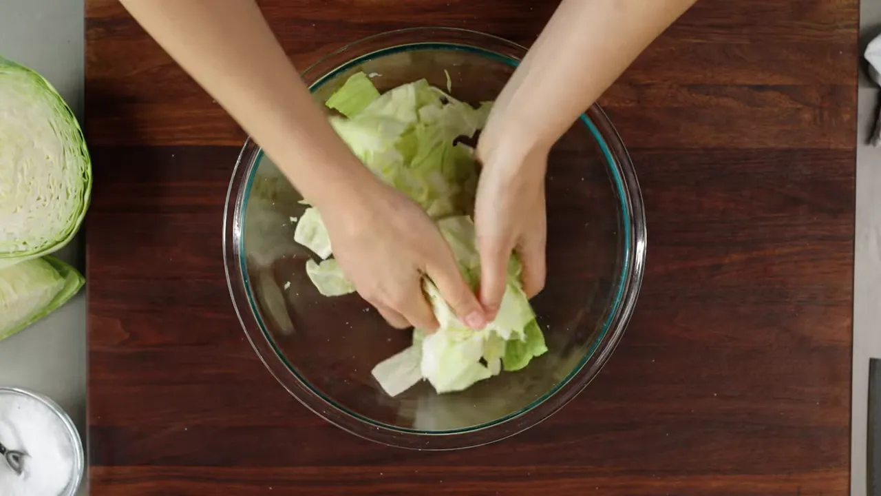 Overhead view of hands massaging cabbage in a glass bowl with halved cabbage and salt nearby