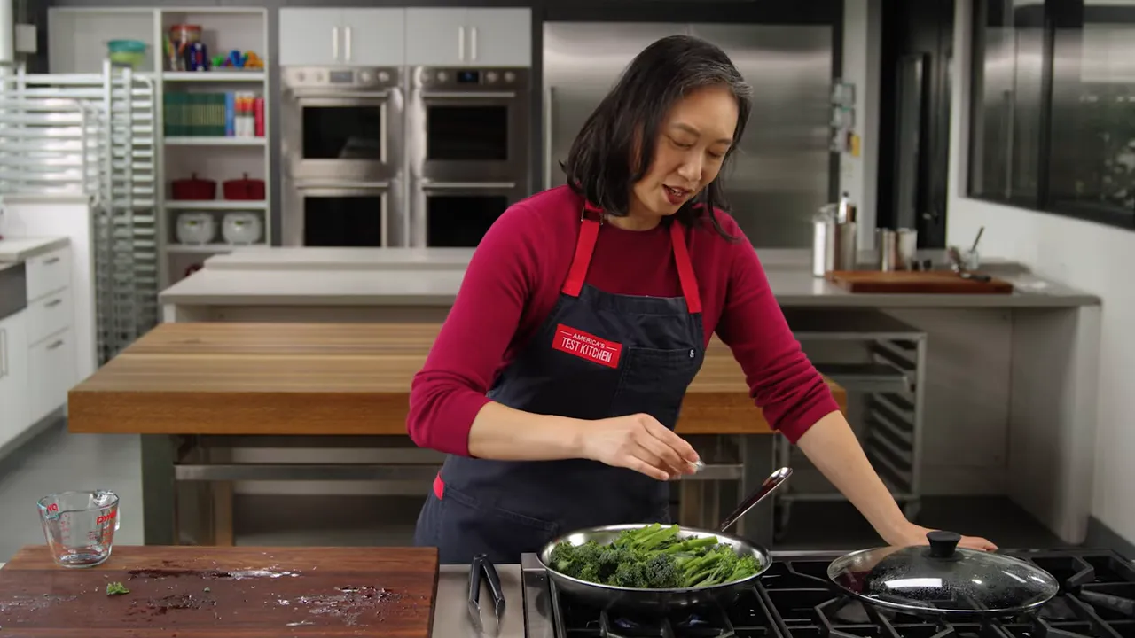 Chef sprinkling seasoning over broccolini simmering in a skillet on the stove.