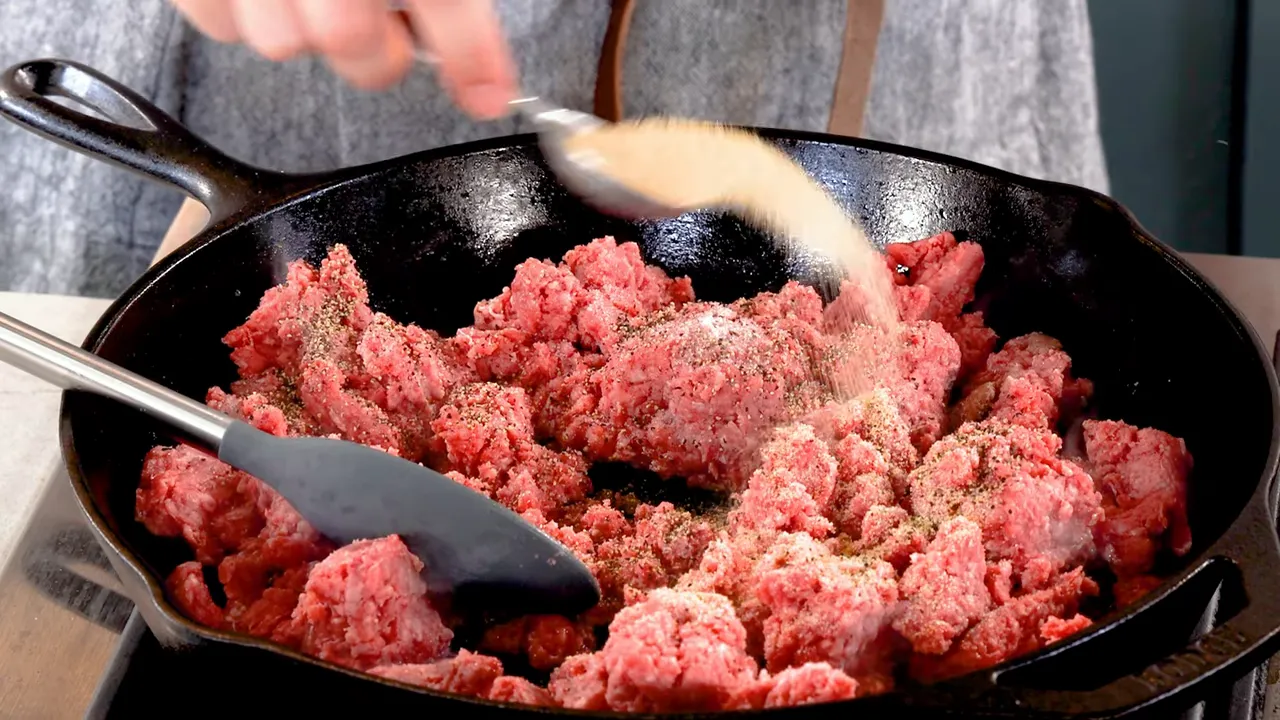 Sprinkling granular seasoning onto ground beef in a hot cast iron skillet