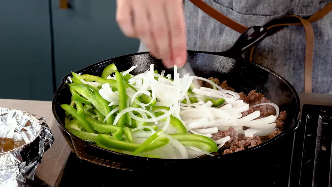 Green bell pepper strips and thinly sliced onions being added to browned ground beef in a cast-iron skillet