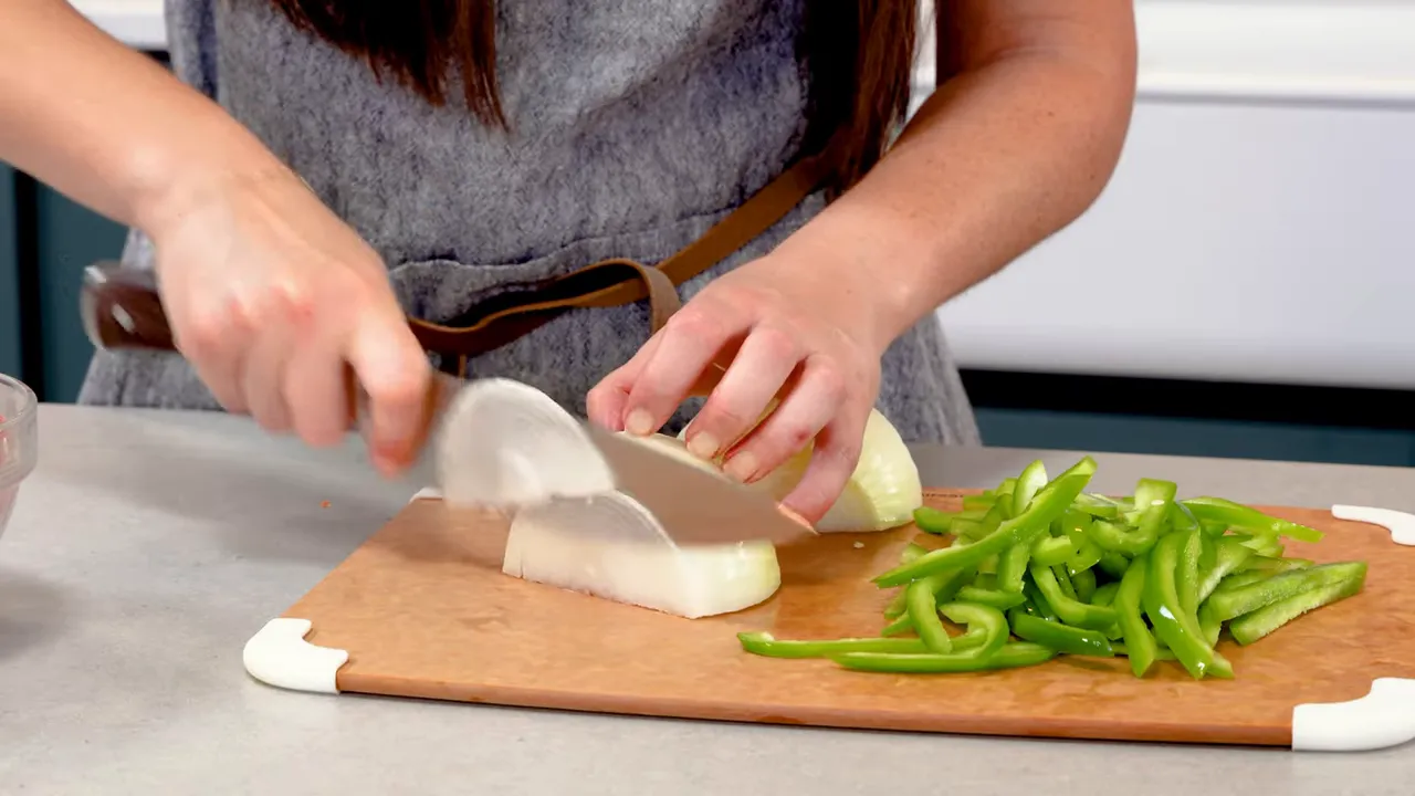 Close-up of hands slicing a yellow onion on a cutting board with a pile of thin green pepper strips beside it