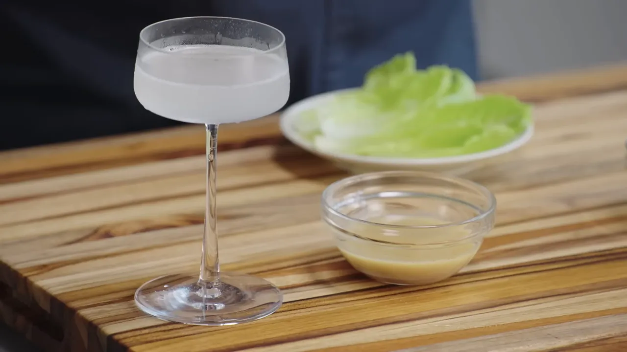 Silky chilled daiquiri in a coupe with a small glass bowl of syrup and a plate of citrus leaves on a wooden board
