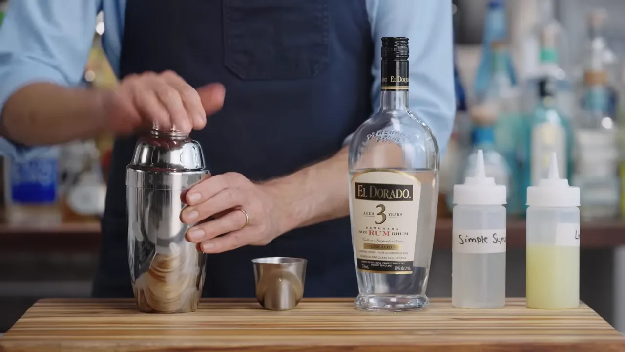 Hands placing the cap on a cocktail shaker next to a bottle of rum and labeled squeeze bottles reading 'Simple Syrup'.