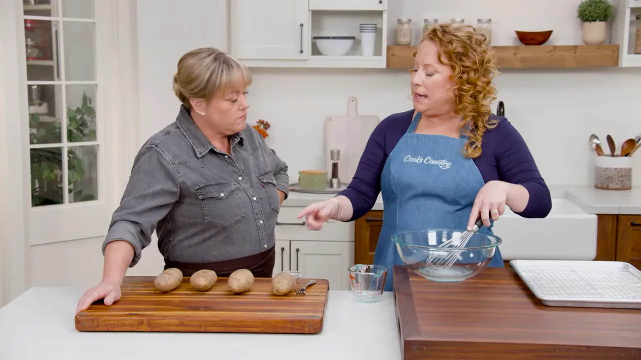 kitchen workstation with four russet potatoes on a cutting board and a mixing bowl with measuring cup on the counter