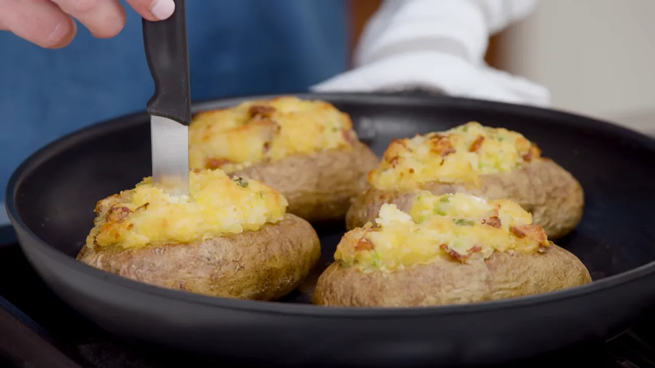 Close-up of four twice-baked potatoes in a skillet with golden, cheesy tops and a knife inserted to show texture.