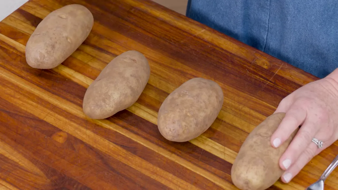 Four russet potatoes on a wooden cutting board with a hand arranging one potato