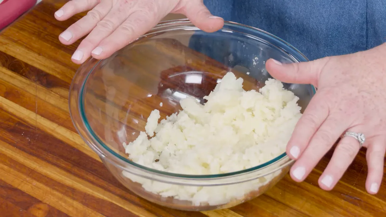 Glass bowl containing scooped potato flesh with hands ready to mix.
