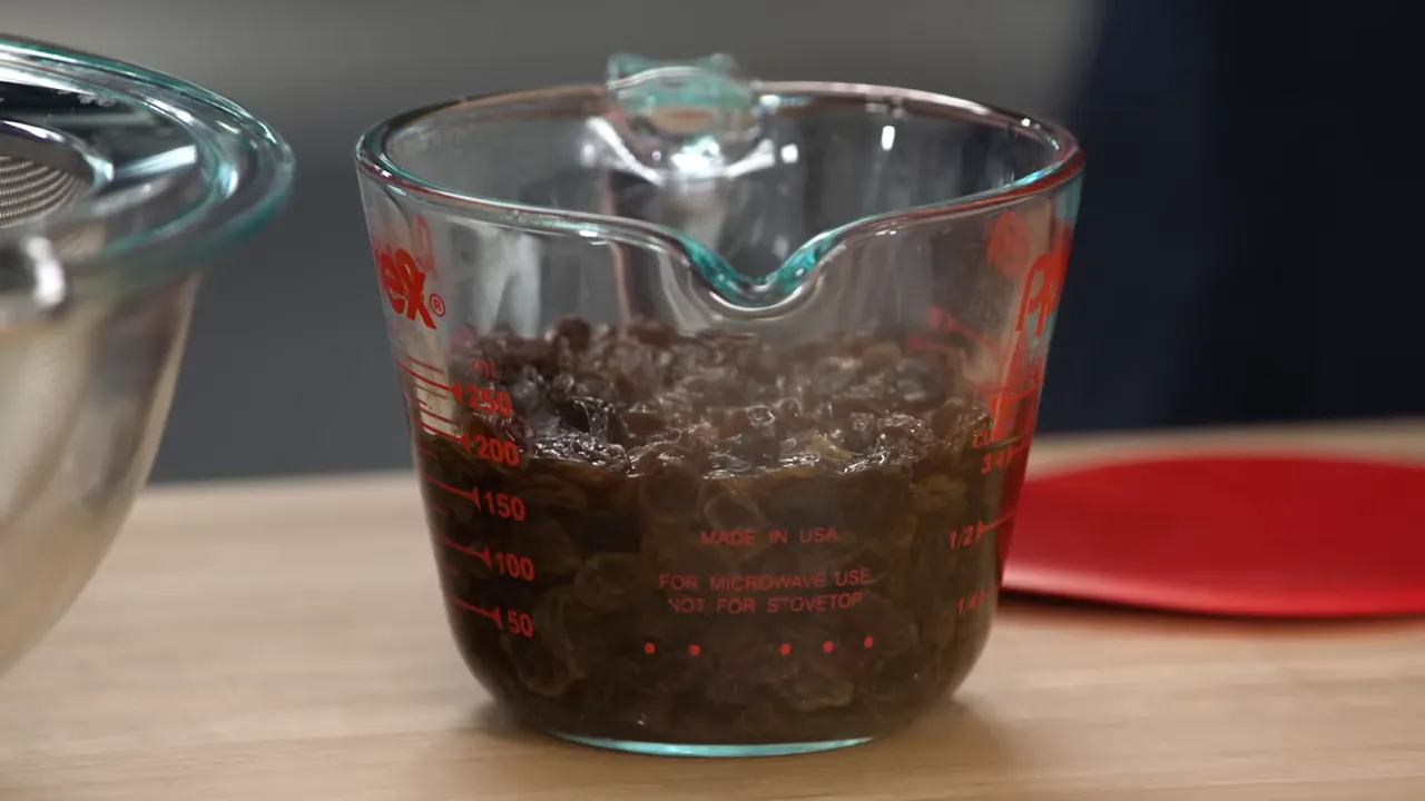 Close-up of a Pyrex measuring cup full of steaming brandy-soaked raisins on a wooden counter