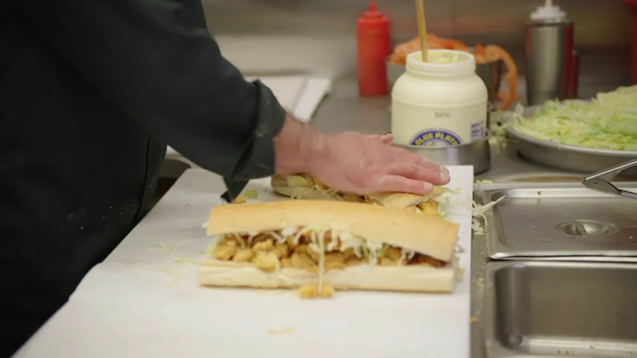 Hand pressing a shrimp po-boy on a counter with jar of mayo and shredded lettuce nearby
