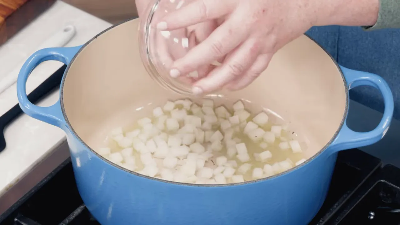 diced onions being poured from a small glass bowl into a blue Dutch oven with oil