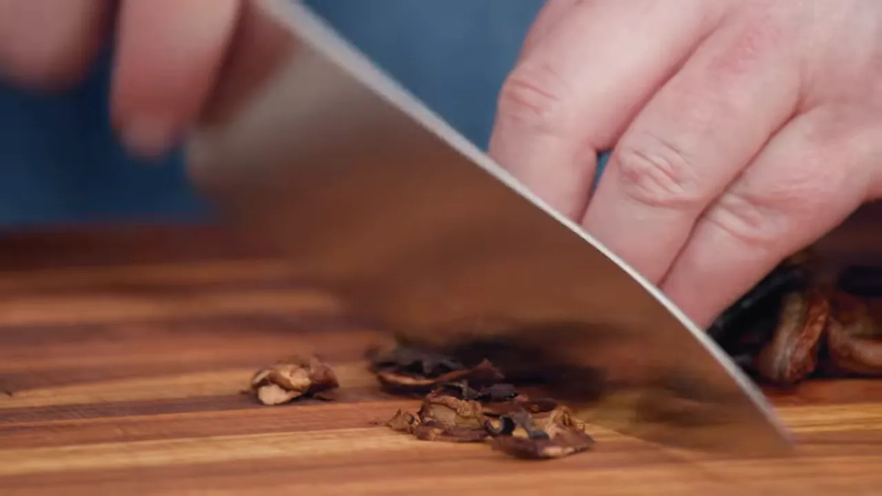 close-up of a cleaver chopping rehydrated porcini mushrooms on a wooden cutting board