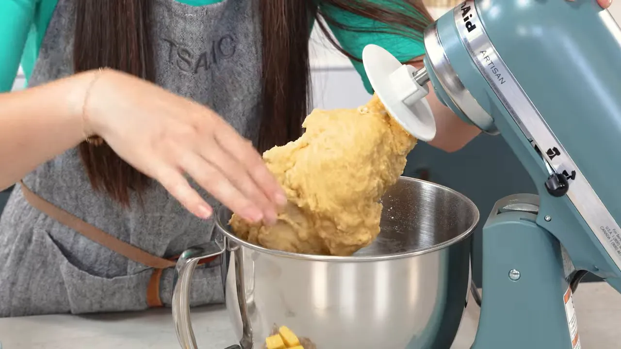 Hands pulling soft, glossy brioche dough from a stand mixer paddle with cubes of butter visible in the bowl.
