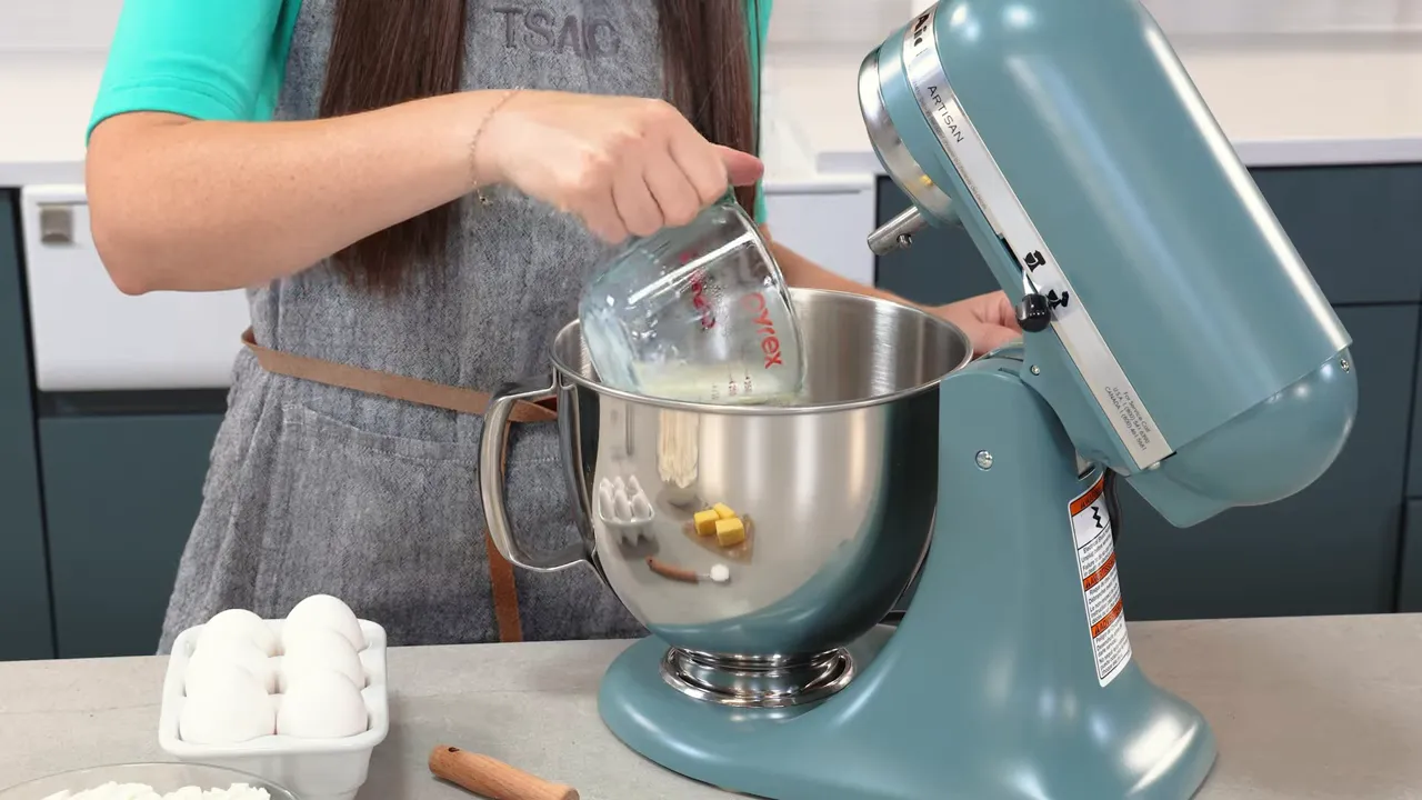 Hand tilting a measuring cup to pour warm milk into a stand mixer bowl with eggs and butter on the counter.