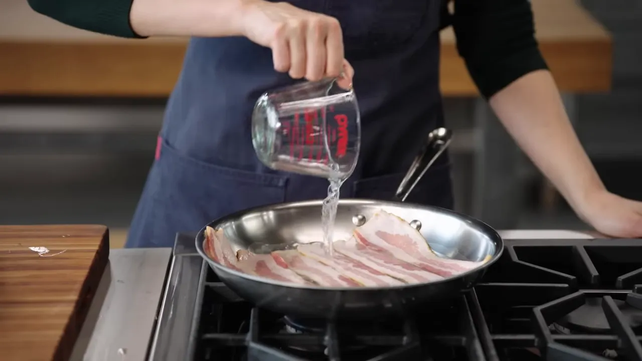 Pouring water from a measuring cup into a skillet of raw bacon to begin a water-simmer.