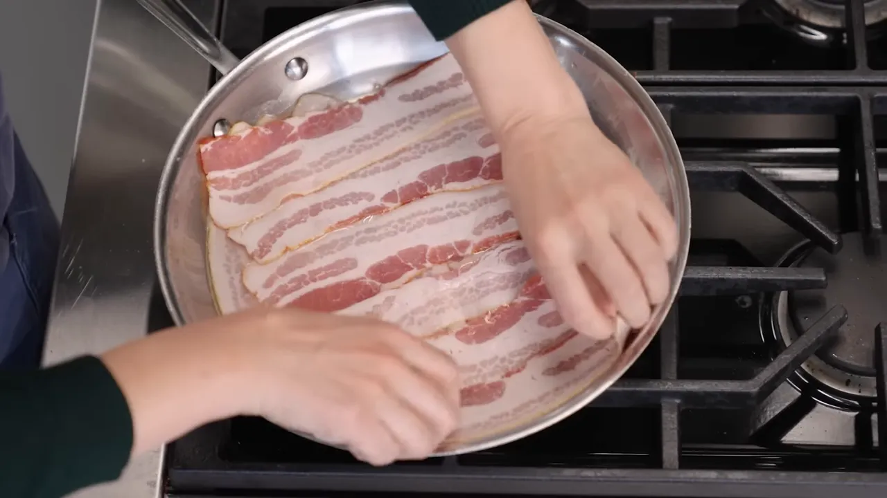 Overhead view of bacon strips laid flat in a stainless skillet with hands adjusting the edges.