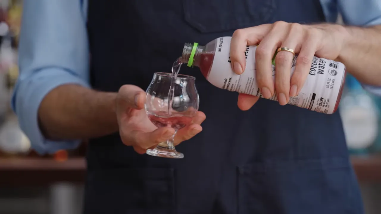 Hands pouring coconut water from a bottle into a small stemmed tasting glass