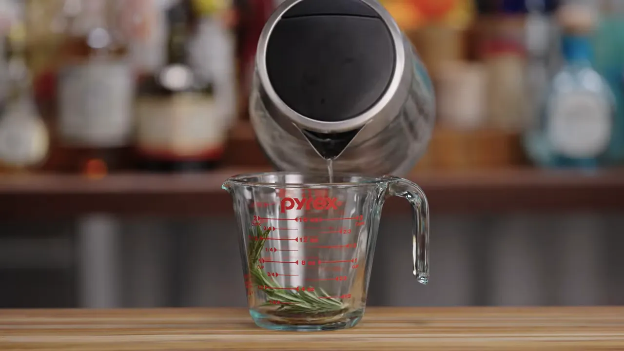 Hot water being poured from a kettle into a Pyrex measuring cup containing tea leaves and a sprig of rosemary