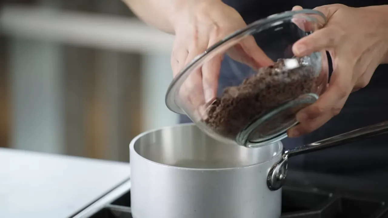 Hands tipping a glass bowl of chopped chocolate into a small saucepan on the stove